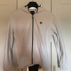 Vit hoodie från Lyle & Scott - Säljer en fräsch vit hoodie från Lyle & Scott med dragkedja och huva. Tröjan har ett broderat gult fågelmärke på bröstet, långa ärmar och praktiska fickor framtill. Perfekt för chill dagar eller när du vill ha en clean look.