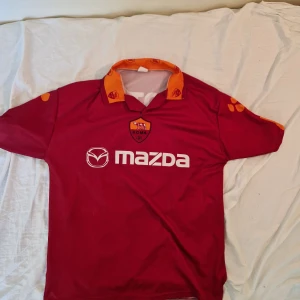 AS Roma Totti 10 röd fotbollströja XL - Säljer en klassisk AS Roma fotbollströja i rött med orange krage och detaljer, Totti 10 på ryggen och Mazda-logga på bröstet. Tröjan är kortärmad och tillverkad i polyester, perfekt för dig som vill ha en ikonisk supportertröja från Italien.