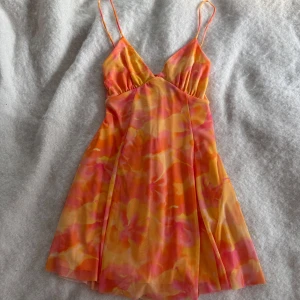 Blommig kort klänning med snörning - Säljer en färgstark kort klänning med blommigt mönster i orange, rosa och gul. Klänningen har tunna axelband, v-ringning och snörning i ryggen för en snygg passform. Materialet är lätt och luftigt, perfekt för varma dagar.