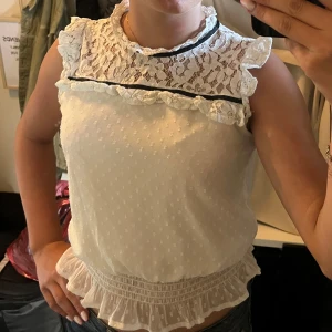 Vit spetsblus med volang från Lindex - Supersöt vit blus från Lindex med spets upptill, volangdetaljer vid axlar och nederkant samt en tunn svart kant vid halsen. Ärmlös modell med lätt transparent tyg och diskret struktur. Perfekt för dig som gillar romantisk och feminin stil.