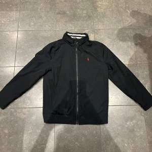 Polo Ralph Lauren jacka - Svart vindjacka från Polo Ralph Lauren med klassisk passform och dragkedja framtill. Jackan har ståkrage, två fickor och den ikoniska röda logon broderad på bröstet. Perfekt för dig som gillar stilren och enkel design. 