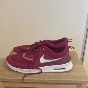 Nike Air Max Thea vinröda sneakers - Nike Air Max Thea sneakers i vinrött med vita swoosh-loggor och detaljer i orange. Skorna har snörning, platt sula och ventilerande mesh på ovansidan. Snygg design med mönstrad hälflik och dämpad sula för extra komfort. Perfekt för dig som gillar sportig stil.