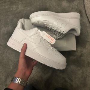 Nike Air Force 1 Low helvita sneakers - Nike Air Force 1 Low i helvitt skinn med klassisk rund tå och platt sula. Snygga detaljer som perforeringar på tån och ikonisk Swoosh-logga på sidan. Skorna har snörning och en clean look som alltid är on point. Perfekta för dig som gillar stilrena sneakers.