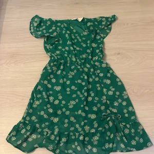 Grön blommig klänning från H&M - Säljer en grön klänning från H&M med sött blommigt mönster och volangkant nedtill. Klänningen har korta ärmar, v-ringning och en fin rosettdetalj på kjolen. Perfekt för sommarens alla tillfällen och riktigt skön att bära.