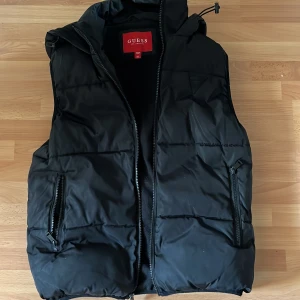 Svart dunväst från Guess med huva - Svart dunväst från Guess med snygg huva och dragkedja framtill. Den är även väldigt varm så man kan ha den på hösten/våren/sommaren. Västen har två praktiska fickor med dragkedja och diskret Guess-logga på bröstet samt broderad logga på huvan. Perfekt för lager-på-lager och chill stil.