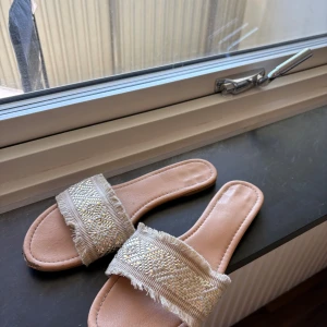 Beige sandaler med broderad detalj - Snygga beige sandaler med bred rem över foten, dekorerad med broderade mönster och små pärlor. Remmen har fransade kanter som ger en chill och bohemisk vibe. Mjuk innersula i syntetmaterial och platt sula för skön komfort. Perfekta för varma dagar.