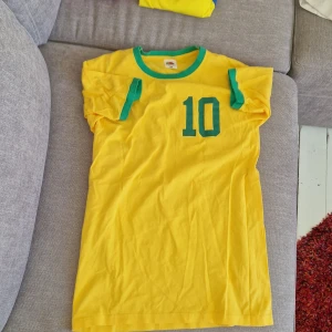 Gul Brasil Pele 10 fotbollströja S - Säljer en klassisk gul Brasilien fotbollströja med gröna detaljer och tryck. Nummer 10 och PELE på ryggen, grönt tryck även fram. Rund hals med grön kant, korta ärmar med gröna ränder. 100% bomull, perfekt för dig som älskar retro och fotboll.