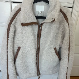 Beige teddyjacka från H&M  - Säljer en beige teddyjacka från H&M i storlek XS. Jackan har bruna detaljer längs dragkedjan, fickorna och ärmarna, samt en hög krage. Materialet är mjuk fleece med syntet och jackan har två fickor framtill och en rejäl dragkedja.