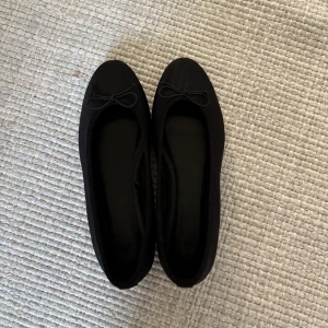 Svarta ballerinaskor med rosett från H&M - Snygga svarta ballerinaskor från H&M med rund tå och en liten rosett framtill. Skorna har en diskret ribbad struktur i tyget och platt sula, vilket gör dem både stilrena och bekväma. Aldrig använda då de är lite för stora!