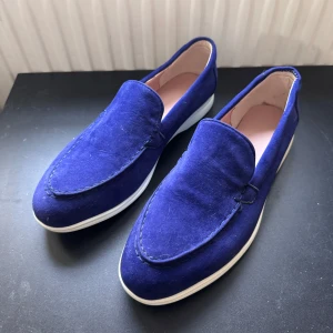 Blå mockaloafers med vit sula - Snygga loafers i djupblå mocka med stilren vit platt sula. Skorna har en klassisk rund tå och är fodrade med ljusrosa insida för extra komfort. Perfekt för dig som vill ha en clean och modern look med lite färg på fötterna.