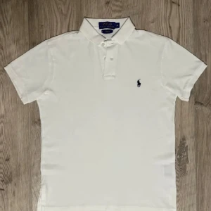 Vit slim fit t-shirt från Polo Ralph Lauren - Vit t-shirt från Polo Ralph Lauren i slim fit med klassisk pikékrage och korta ärmar. Broderad marinblå logga på bröstet. Tillverkad i mjuk bomull som känns skön mot huden. Perfekt för dig som gillar stilrena och tidlösa plagg.
