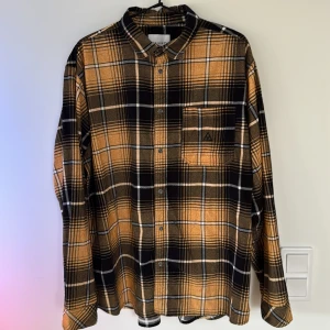 Rutig skjorta från Vailent XL - Snygg rutig skjorta från Vailent i storlek XL. Skjortan har en klassisk krage, bröstficka och knappar framtill. Färgerna är svart, beige och prange med ett tydligt rutmönster. Perfekt för dig som gillar en avslappnad och cool stil.