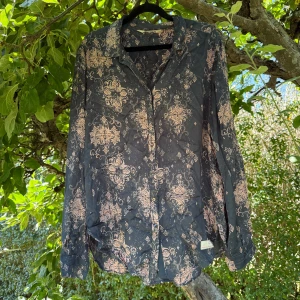 Mörkblå skjorta med rosa mönster - Snygg mörkblå skjorta med unikt rosa mönster som ger en cool vibe. Skjortan har klassisk krage, knappar framtill och långa ärmar. Materialet känns lätt och skönt mot huden, perfekt för dig som gillar att sticka ut med detaljer. Passar dig som vill ha något annorlunda i garderoben. Från märket odd molly. Storlek 3 som enligt deras storleksguide motsvarar en storlek L.  Mycket fint skick, jag hittar inga synliga anmärkningar eller defekter.