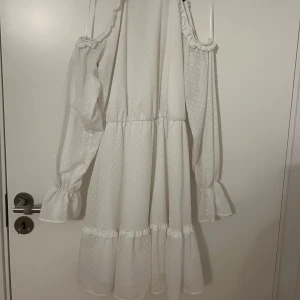 Vit prickig klänning från Mohito - Superfin vit långklänning från Mohito med små prickar i tyget. Klänningen har offshoulder och långa ärmar med volangdetaljer vid axlar och ärmslut. Den har knytning i nacken och ett luftigt, flowy fall som ger en romantisk vibe. Perfekt för sommarens alla tillfällen! Den är använd bara en gång.