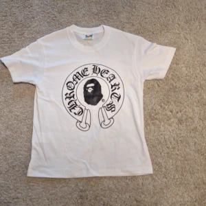 Vit BAPE x Chrome Hearts t-shirt S - Vit t-shirt från BAPE x Chrome Hearts med stor svart print på både fram- och baksidan. Framsidan har Chrome Hearts-logga och BAPE-ansikte, baksidan har Baby Milo, Chrome Hearts-kors och text. Klassisk rund hals och korta ärmar. Tillverkad i mjuk bomull.