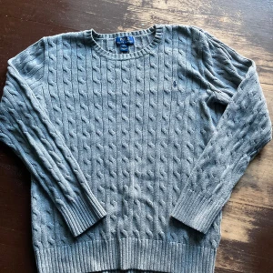 Kabelstickad Ralph Lauren Tröja - Snygg grå kabelstickad tröja från Polo Ralph Lauren med klassisk rund hals och diskret broderad logga på bröstet. Tröjan har långa ärmar och ribbade muddar vid ärmslut och nederkant. Perfekt för dig som gillar stilrena och tidlösa plagg.