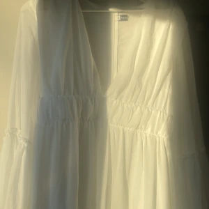 Vit omlottklänning från Miss - Säljer en vit omlottklänning från Miss med v-ringning och långa, transparenta ärmar. Klänningen har volanger och smockdetaljer i midjan och på ärmarna, vilket ger en flowy och romantisk vibe. Liten i storleken! Perfekt för sommarens alla tillfällen!