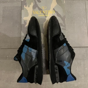 Valentino Garavani Rockrunner sneakers - Snygga Valentino Garavani Rockrunner sneakers med svart och blått kamouflagemönster. Skorna har svarta snören, platt sula och detaljer i mocka och skinn. Ikoniska nitar på sidan och blå häl ger extra edge. Perfekt för dig som gillar snygg stil och exclusive designer 😁😁 (små skador på skon)
