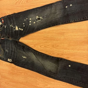 Dsquared2 mörka slim jeans med detaljer - Säljer ett par mörkblå Dsquared2 jeans i slim fit med coola slitningar och vita färgstänk på både fram- och baksida. Jeansen har klassiska fem fickor, kontrastsömmar och en snygg Dsquared2-tag vid fickan. Perfekt för dig som gillar streetstyle och unika detaljer.