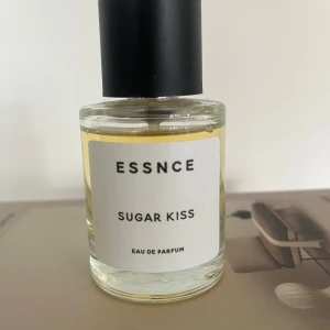 ESSNCE Sugar Kiss Eau de Parfum - Parfymen Sugar Kiss från ESSNCE kommer i en rund, transparent glasflaska med svart cylinderformad kork. Vätskan är ljusgul och etiketten är vit med svart text. Modern och stilren design som känns fräsch och ungdomlig.