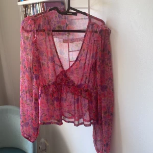 Blommig volangblus från BikBok - Superfin transparent blus från BikBok i rosa och lila blommigt mönster. Blusen har v-ringning, volangdetaljer och långa ärmar. Perfekt för dig som gillar romantisk och färgglad stil. Luftigt material som känns lätt och flowy.