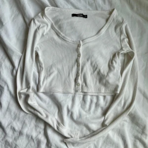 Vit croppad ribbad topp från Bik Bok - Superfin vit croppad topp från Bik Bok med långa ärmar och ribbad struktur. Toppen har knappar framtill och en snygg, djup rundad halsringning. Perfekt att styla med höga jeans eller kjol för en clean look.