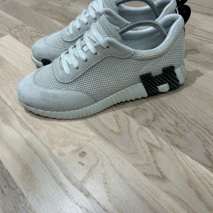 H vita sneakers med mesh - Säljer ett par stilrena vita sneakers från H med cool meshpanel och mockadetaljer. Sulan har en unik design med svarta och vita inslag som sticker ut. Skorna har snörning och en platt sula, perfekt för dig som gillar minimalistisk och modern stil.