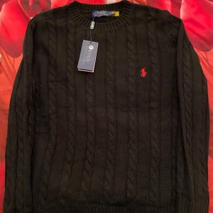 Svart kabelstickad tröja Polo Ralph Lauren - Svart kabelstickad tröja från Polo Ralph Lauren i storlek M. Klassisk rund hals och långärmad modell med röd broderad logga på bröstet. Tröjan är gjord i mjuk bomull och har ribbade muddar vid hals, ärmslut och nederkant. Perfekt för dig som gillar stilrena och tidlösa plagg.