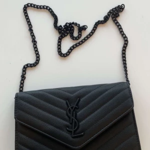 YSL väska - Snygg svart kuvertväska från YSL med kedjerem och ikonisk logga framtill. Väskan har quiltad design i chevronmönster och är tillverkad i skinn. Perfekt för att lyfta din outfit med en lyxig touch.