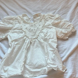 Vit blus med spets från Gina Tricot - Supersöt vit blus från Gina Tricot med puffiga korta ärmar och snygg spetsdetalj framtill. Blusen är gjord i 100% bomull och har en luftig passform. Perfekt för dig som gillar romantiska och trendiga vibes. Passar dig som vill ha något extra till din outfit.