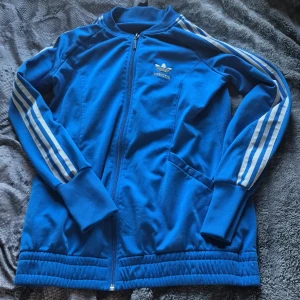 Blå Adidas track jacket strl 38 - Säljer en blå Adidas track jacket med klassiska vita ränder längs ärmarna och Adidas logga på bröstet och ryggen. Jackan har dragkedja framtill, ribbade muddar och sidofickor. Perfekt för dig som gillar sportig och retro stil. Original priset är 600kr.