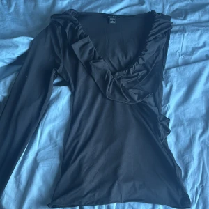 Svart volangblus från Amisu - Svart blus från Amisu i storlek S med lång ärm och djup v-ringning. Snygga volangdetaljer längs halsringningen och framtill ger en cool och trendig look. Mjukt och stretchigt material som sitter bekvämt och följer kroppen.