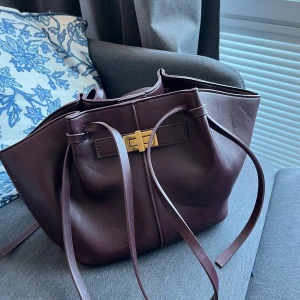 Vinröd handväska från Gina Tricot 💕 - Snygg vinröd handväska i skinnimitation från Gina Tricot med guldfärgat vridlås och långa snörningsdetaljer. Väskan har rymligt innerfack och en stilren, mjuk form som gör den enkel att matcha med olika outfits. Perfekt storlek för allt du behöver ha med dig. Den är i bra skick. Long strap midi bag från Gina Tricot 💕