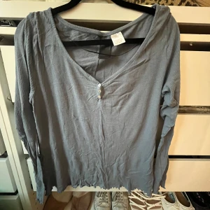 Grå ribbad långärmad tröja - Säljer en chill grå långärmad tröja från H&M med ribbad struktur och v-ringning med tre små knappar. Tröjan har en lite lös passform och rå kant nedtill för en avslappnad vibe. Perfekt att slänga på sig när man vill vara bekväm men ändå se snygg ut.