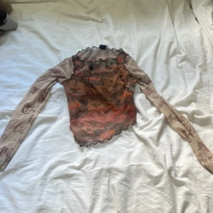 Mesh topp från Urban Outfitters XS - Säljer en unik mesh topp från Urban Outfitters i storlek XS. Toppen har långa ärmar med tryckt text, asymmetrisk nederkant och volangdetaljer. Färgerna går i beige, brunt och orange med ett coolt mönster på kroppen. Perfekt för dig som gillar statement-plagg!