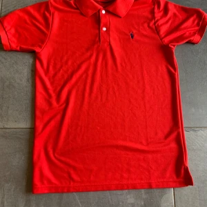 Polo Ralph lauren piké  - Snygg röd polo t-shirt från Ralph Lauren med klassisk krage och två knappar framtill. T-shirten har korta ärmar och den ikoniska blå Polo-loggan broderad på bröstet. Tillverkad i mjuk bomull som är skön mot huden. Perfekt för en chill och stilren look. Storleken är M och neck tagen är bort klippt och därav priset 