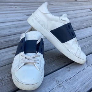 Valentino Garavani vita sneakers - Säljer ett par snygga Valentino Garavani sneakers i vitt skinn med bred marinblå rem över vristen och coola nitar på hälen. Skorna har rund tå, platt sula och klassisk snörning. Perfekt för dig som gillar exklusiv streetstyle och vill sticka ut med designer på fötterna.