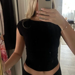 Svart topp med öppen rygg - Säljer en svart, croppad topp med korta ärmar och riktigt snyggt öppen rygg. Toppen har en tight passform och är perfekt om du vill ha en enkel men ändå cool look. Materialet känns mjukt och stretchigt, så den sitter skönt mot kroppen.