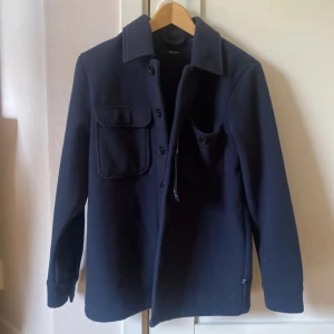 Mörkblå overshirt från Matinique XS - Snygg mörkblå overshirt från Matinique i storlek XS. Jackan har klassisk krage, två stora bröstfickor med knappar och knäppning hela vägen framtill. Materialet är mjukt och känns som ull, perfekt för lager på lager. Stilren och enkel design som funkar till många outfits.