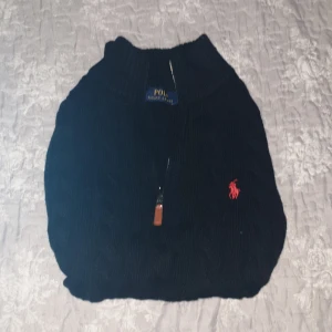 Svart stickad tröja Polo Ralph Lauren - Svart kabelstickad tröja från Polo Ralph Lauren i storlek M. Tröjan har en half zip-dragkedja framtill och den klassiska röda Polo-loggan broderad på bröstet. Mjuk och varm, perfekt för kyliga dagar. Långärmad och med ribbade muddar för extra komfort. helt ny!