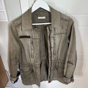 Grön jeansjacka från Wera Stockholm - Snygg beige parkajacka från Wera Stockholm med dragkedja och knappar framtill, flera fickor och justerbar midja. Jackan har långa ärmar och coola detaljer som ger en avslappnad men stilren look. Perfekt för dig som gillar streetstyle.