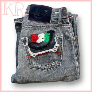 Jacob Cohen jeans - Unika jeans från Jacob Cohen i klassisk grå  tvätt med röda kontrastsömmar och broderad bakficka i rött, vitt och grönt. Modellen har rak passform, knappgylf och coola detaljer som fransad fickkant och specialbroderi. Perfekt för dig som vill sticka ut.