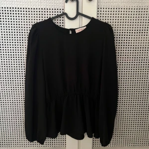 Svart blus med volang - Säljer en svart blus från Only med rund halsringning och volang nedtill. Blusen har långa ärmar med lätt puff och knäppning bak i nacken. Perfekt att styla med jeans eller kjol för en clean look.