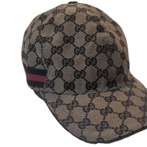 Gucci beige keps med GG-mönster - Snygg keps från Gucci i beige med klassiskt GG-monogram över hela kepsen och en svart/röd rand på sidan. Kepsen har böjd skärm och justerbar rem baktill. Tillverkad i slitstark bomull med insida i mörkblått tyg. Perfekt för dig som gillar lyxiga accessoarer.