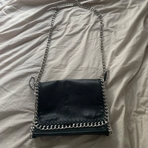 Svart axelväska med kedja från TIAMO - Snygg svart axelväska från TIAMO i skinnimitation med  silverkedja som axelrem och runt kanten. Väskan har magnetlås, innerfack med dragkedja och plats för småsaker. Perfekt storlek för det viktigaste och ger en cool vibe till din outfit. Pris kan sänkas vid förhandling. 