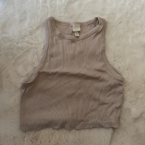 Beige ribbad croppad linnetopp H&M - Säljer en beige croppad linnetopp från H&M i ribbad bomull. Toppen är ärmlös med hög hals och har en clean, enkel look som är lätt att matcha med jeans eller kjol. Perfekt för dig som gillar minimalistisk stil och vill ha en topp som känns både chill och snygg.