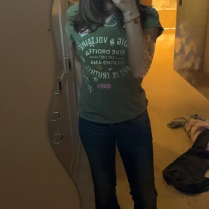 Grön t-shirt från Zadig & Voltaire - Cool grön t-shirt från Zadig & Voltaire med tryck i vitt, rosa och gult. T-shirten har korta ärmar och rund hals. Trycket har blommor, hjärtan och stjärnor som ger en lekfull vibe. Perfekt för dig som gillar statement-plagg och snygga detaljer.