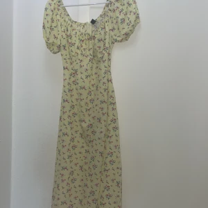 Blommig ljusgul långklänning - Säljer en söt medellång klänning i ljusgul färg med blommigt mönster i rosa och grönt. Klänningen har puffiga korta ärmar, rundad halsringning med rynk och en slits framtill. Perfekt för sommarens alla tillfällen!