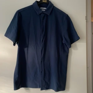 Mörkblå kortärmad skjorta från Zara - Snygg mörkblå skjorta från Zara i regular fit med klassisk krage och korta ärmar. Skjortan har knappar framtill och är tillverkad i ett mjukt bomullsmaterial som känns skönt mot huden. Perfekt för dig som gillar stilrena och enkla plagg.