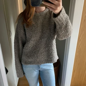 Grå stickad tröja - Mysig grå stickad tröja med rund hals och lång ärm. Tröjan har en loose passform och är tillverkad i ett fluffigt, strukturerat material som känns extra skönt under kyliga dagar. Perfekt att slänga på sig när man vill vara både bekväm och snygg.